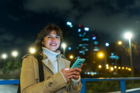 Woman standing on a city bridge at night, smiling and typing on her mobile phone, with illuminated
