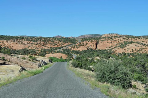A road leads through a dry landscape with rocks and scattered greenery under a blue sky, Grand