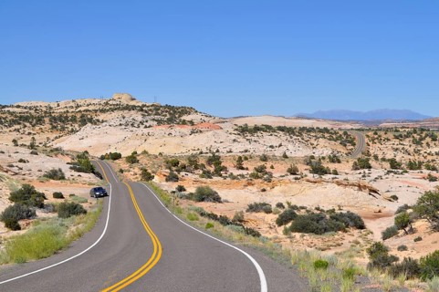 Asphalt road leads through dry, hilly landscape with reddish rocks and vegetation under blue sky,