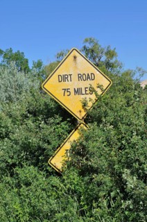 A yellow road sign with the words 'Dirt Road 75 Miles' appears from lush vegetation, Grand