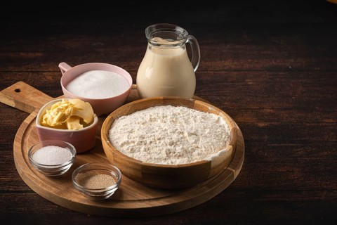 Baking ingredients on a wooden table with a bowl of flour, sugar, and a pot of milk