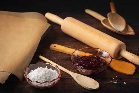 Rolling pin, cooking spoon and baking paper next to jam and sugar on a dark wooden table in a