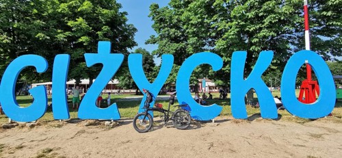 A bicycle stands in front of a large blue lettering of 'Gizycko' on a sandy beach, Masuria bicycle