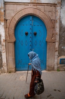 Tunis, Tunisia. 7th May 2024 An old woman wearing a blue shawl walks past a traditional blue door