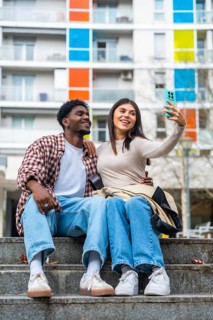 Happy diverse young couple sitting on concrete steps, smiling and taking a cheerful selfie together