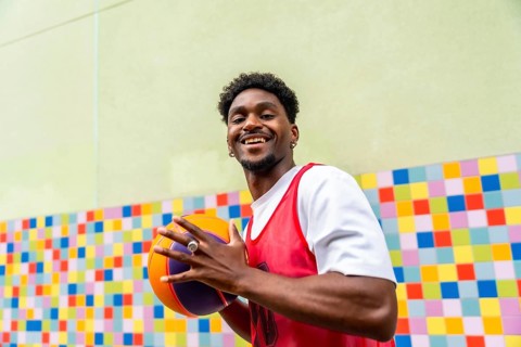 Young man smiling and holding a vibrant basketball in sportswear, standing against a colorful tiled