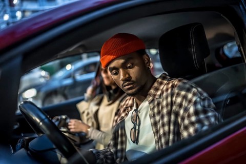 Young man wearing a beanie and plaid shirt sitting in a parked car with a woman, looking down with