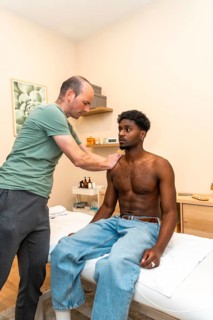 Physical therapist massaging a young black man's shoulder while he sits on an examination table,