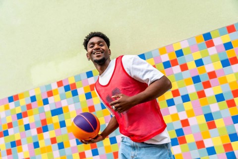 Young black man happily holding a basketball, dressed in a red sports vest and jeans, posing