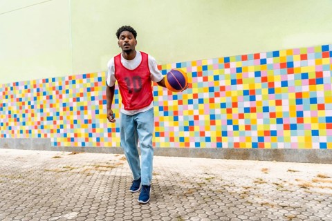 Young black man in casual sportswear playing and dribbling a basketball on a paved court against a