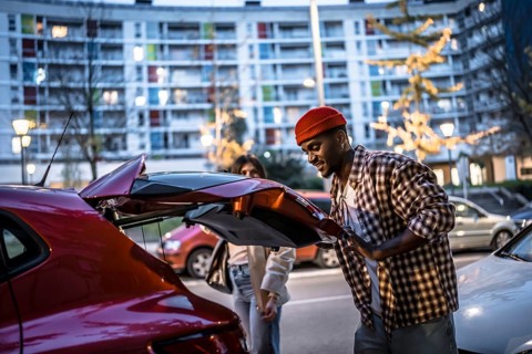 Young couple standing by their car with an open trunk in a city street at dusk, preparing for a