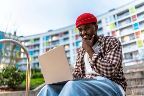 Young black man sitting on apartment steps with laptop, smiling and thoughtful as he works or