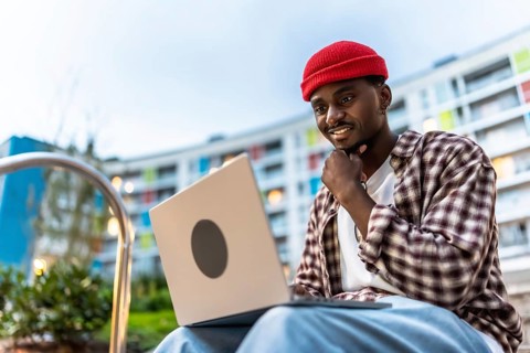 Young black man smiling while working on a laptop computer with casual clothes and a red beanie,