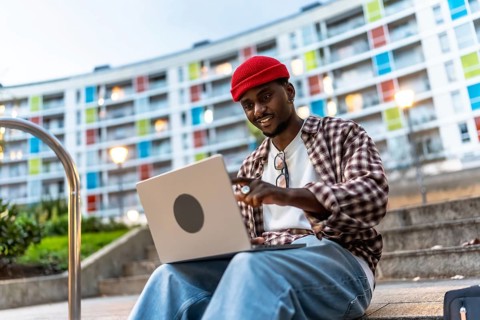 Young black man in casual clothing using a laptop while sitting on steps in an urban setting,