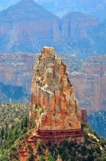 A massive rock rises steeply in a canyon landscape, surrounded by other rocks and sparse