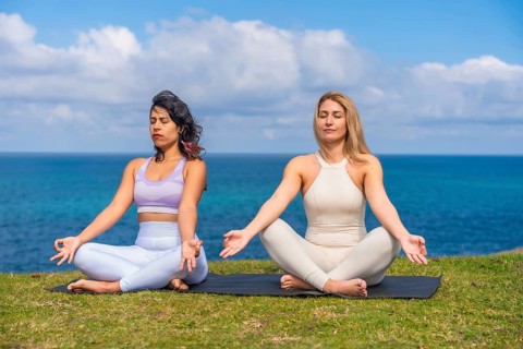 Two women sit cross legged on a mat by the ocean, practicing yoga and meditation together at
