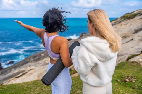Two women standing together on a grassy cliff overlooking the ocean, one raising an arm and holding