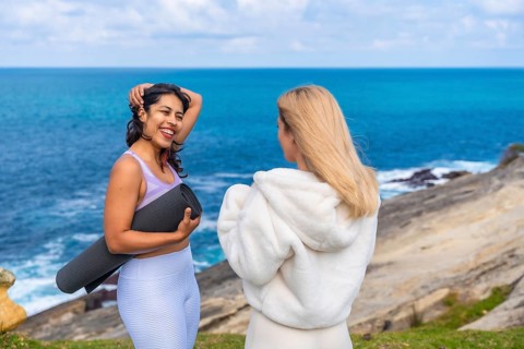 Two women friends standing on a cliff overlooking the ocean, one holding a yoga mat and laughing
