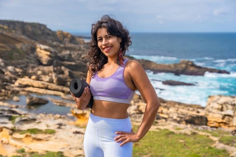 Woman standing on a rocky coastline holding a yoga mat, preparing for an outdoor fitness session