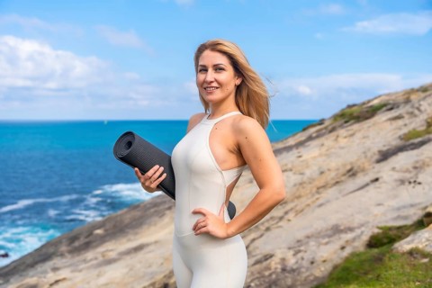 Woman standing confidently on a coastal cliff with a yoga mat, embracing a healthy and balanced