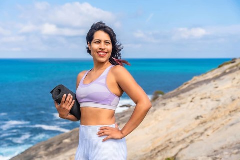 Woman smiling, holding a rolled yoga mat on a rocky cliff, looking towards the bright blue ocean,