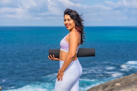 Woman smiling and holding a black yoga mat, standing on a cliff edge with the vast blue ocean and