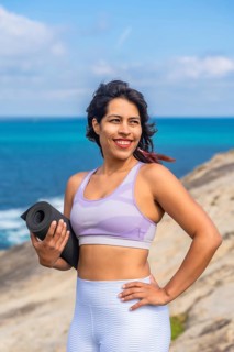Woman holding a yoga mat and smiling, standing on a rocky cliff overlooking the blue ocean during