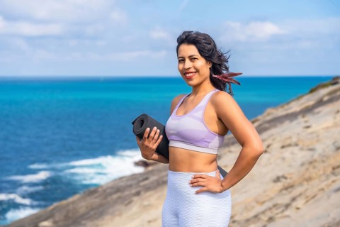 Woman smiling and holding a rolled yoga mat, standing confidently on a coastal cliff overlooking
