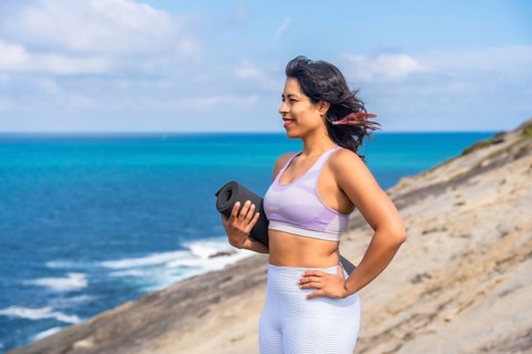 Woman standing on cliff by the ocean, holding a yoga mat and looking to the side, embodying
