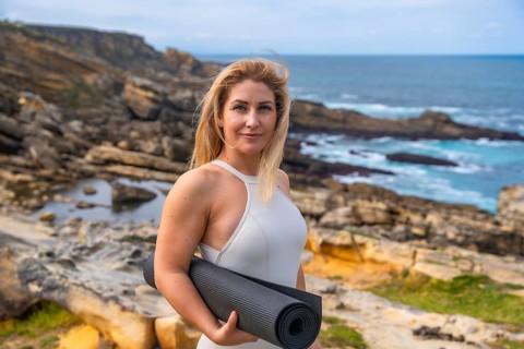 Blonde woman holding a yoga mat on a rocky coast, standing and preparing for outdoor meditation and