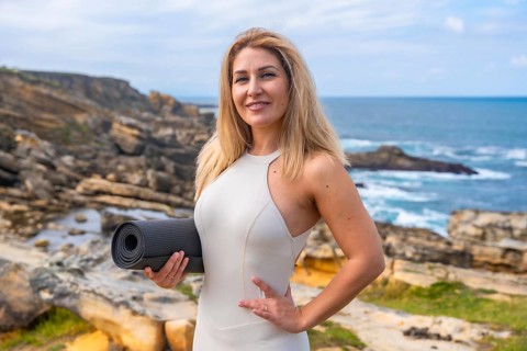 Woman stands on rocky coast by the ocean holding a yoga mat, smiling at camera before a yoga and