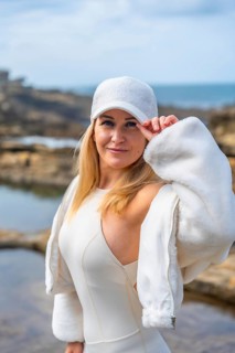 Young woman wearing a white cap and dress, standing confidently on rocky coastal terrain, with the