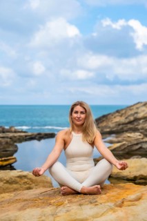 Woman in a white outfit sitting in a lotus position on a rock by the ocean, meditating and finding