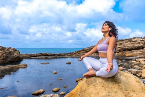 Woman in sportswear practicing yoga and meditation in lotus position on a large rock by the sea,
