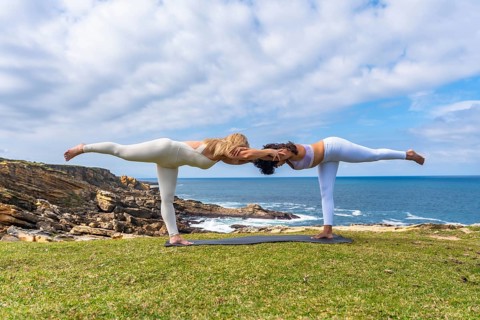 Two women practicing acro yoga outdoors on a grassy cliff overlooking the ocean, holding a deep