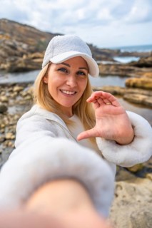 Blonde woman in white cap and jacket smiling at camera on a rocky beach, forming a heart with her