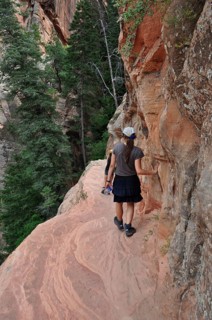 Hiker on a narrow trail to Angel's Landing viewpoint along red rocks surrounded by green trees,