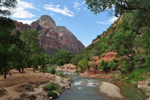 A river flows through a mountainous landscape with green trees and cliffs under a blue sky, Zion