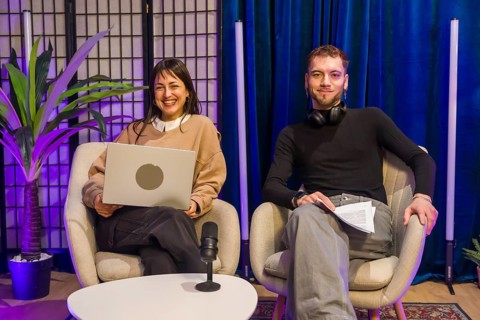 Two smiling content creators, a woman holding a laptop and a man holding notes, sitting in chairs