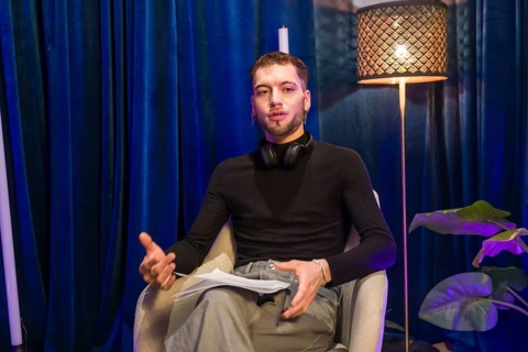 Young man wearing headphones around his neck, holding papers, and gesturing while speaking,