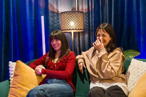 Two young women sitting on a sofa, one smiling and the other laughing, collaborating creatively in