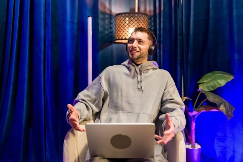 Young man wearing headphones and hoodie sitting with open laptop, actively speaking and gesturing