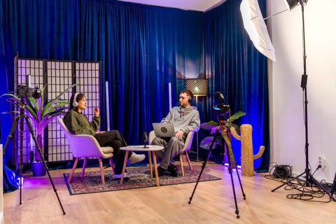 Two young adults in a professional podcast studio record an interview with headphones, laptop,
