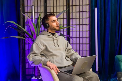 Young man wearing headphones and holding a laptop, talking and recording content for a podcast or