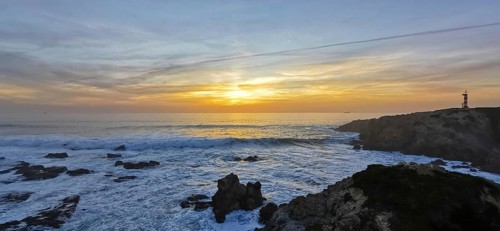A lighthouse watches over the turbulent sea at sunset, the waves hit the rocks, hike the