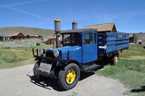 A blue historic truck with yellow wheels, Dodge Graham 1927, stands in front of a wooden house
