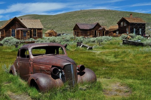 Rusty car wreck, Chevrolet Coupé, on green field in abandoned town with wooden houses, Bodie Ghost