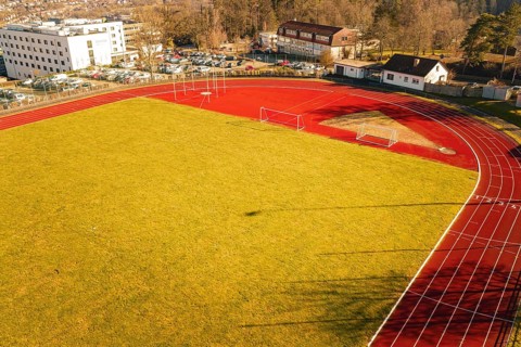 Aerial view of a sports field with red running track, yellow lawn and surrounding buildings,