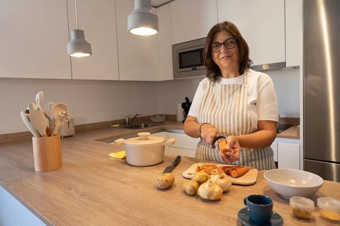 Smiling mature woman wearing apron peeling carrot while preparing vegetables in modern kitchen.