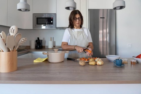 Mature woman wearing apron peeling carrot while preparing vegetables in modern kitchen. Healthy